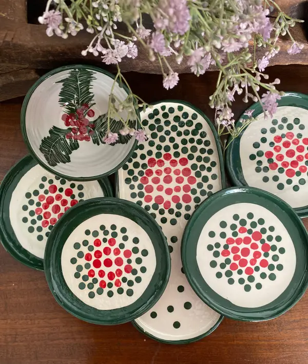 White ceramic plates and bowl with dark green and red dot patterns and holly illustration, styled with purple flowers.