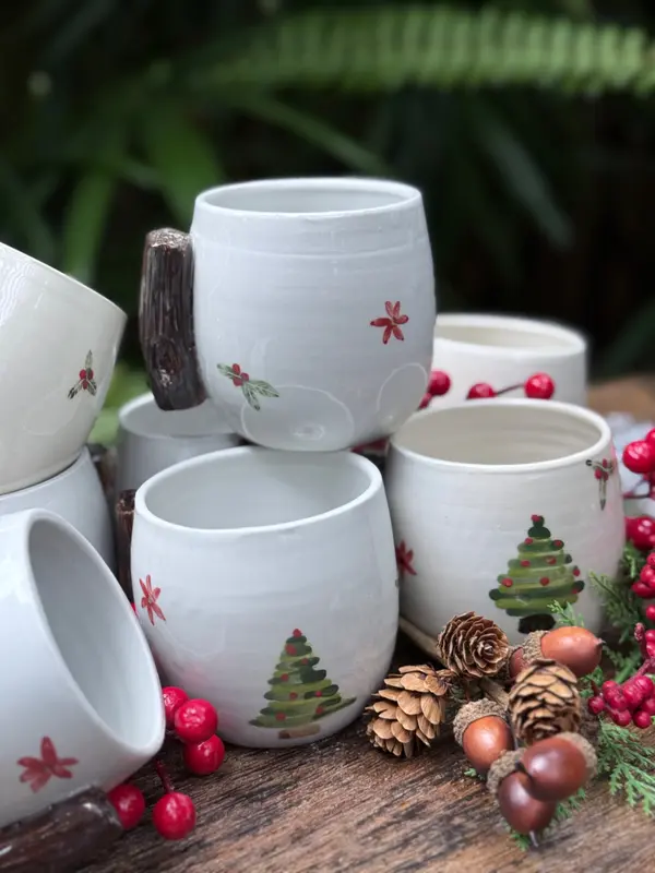 White ceramic mugs with hand-painted Christmas trees and holly berries, featuring textured wood-style handles.