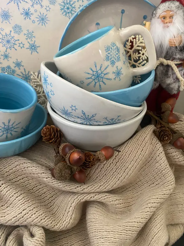 White and light blue ceramic bowls and mug featuring snowflake designs styled with pine cones and winter accents.