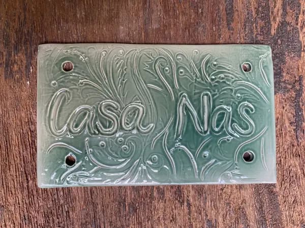 Sage green ceramic wall plaque with 'Casa Nas' in raised script and intricate swirled patterns.