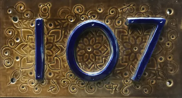 Ceramic house number plaque with cobalt blue numbers 107 on a brown textured floral background.