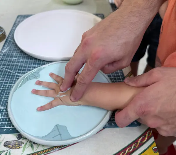Adult hands guiding a child's hand into a light blue glazed ceramic plate to create a handprint impression.