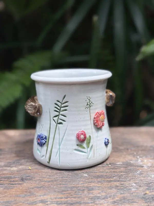 White ceramic vase with green carved stems and small colorful 3D flowers on a rustic wooden background.