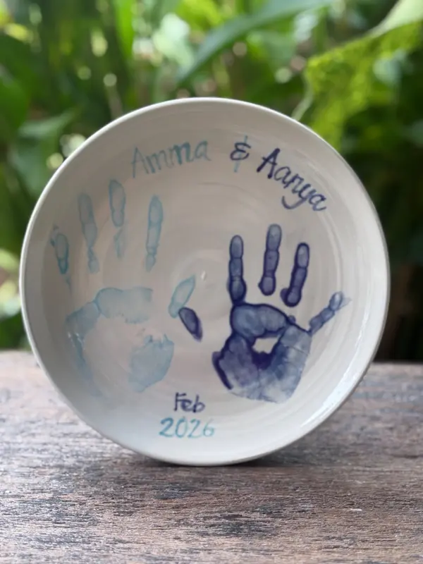 White ceramic bowl with two blue handprints, handwritten names, and a date.