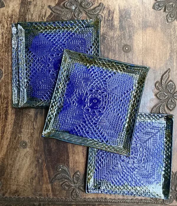 Three square ceramic plates with cobalt blue lace textures and dark metallic rims on a carved wood table.