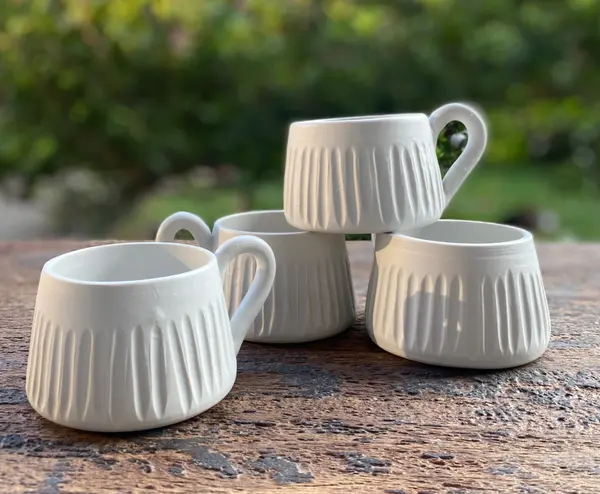 Four matte white mugs with vertical fluted carvings on a rustic wooden surface against a garden background.