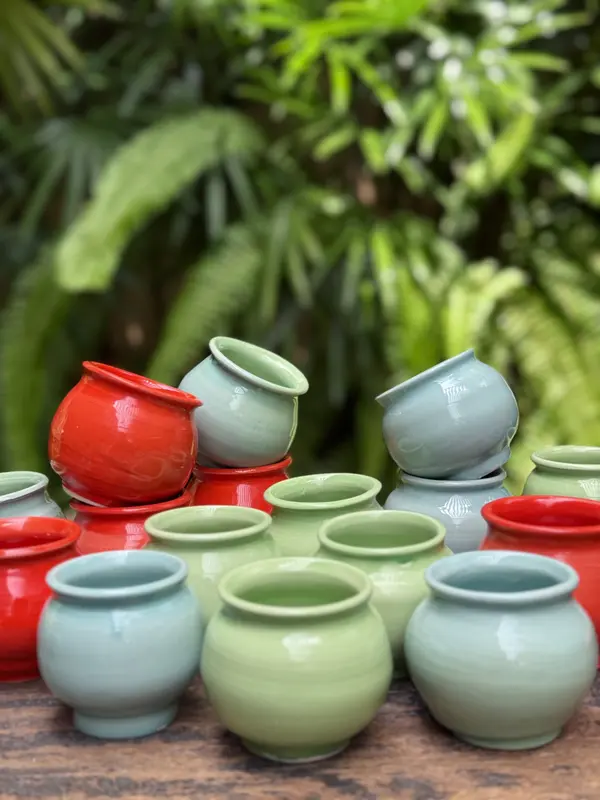Small colorful ceramic jars in red, light green, and light blue glazes against a blurred leafy background.