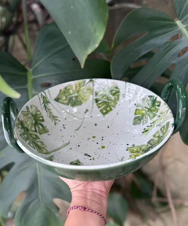 White ceramic bowl with green monstera leaf patterns, dark green handles and exterior, held against leafy background.