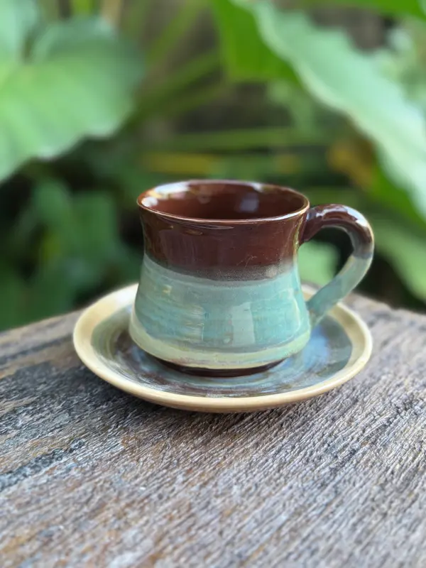 Handmade ceramic mug and saucer set with dark brown and turquoise glazes on a rustic wood surface.