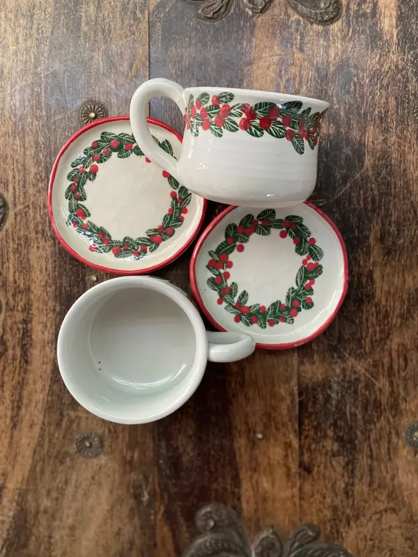 White ceramic mugs and plates featuring hand-painted green leaves and red berry wreath patterns on a wood background.