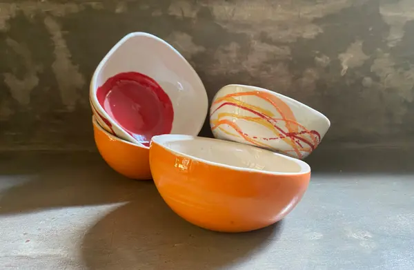 Four colorful ceramic bowls in orange, red, and white with abstract painted lines on a concrete surface.