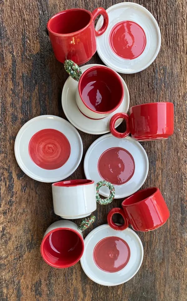 Red and white ceramic mugs and saucers with patterned handles on a rustic wood surface.