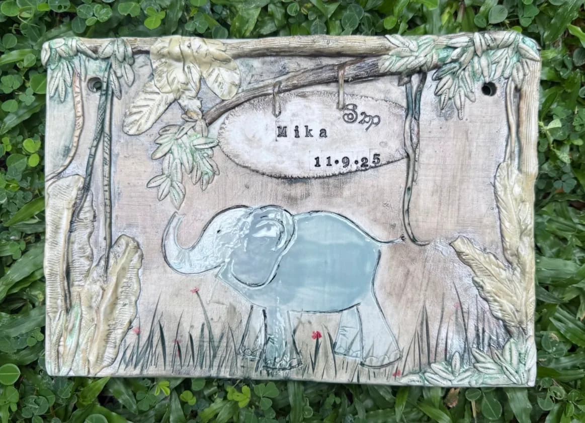 Ceramic wall plaque with a blue elephant relief, jungle leaves, and the name Mika 11.9.25 on a beige background.