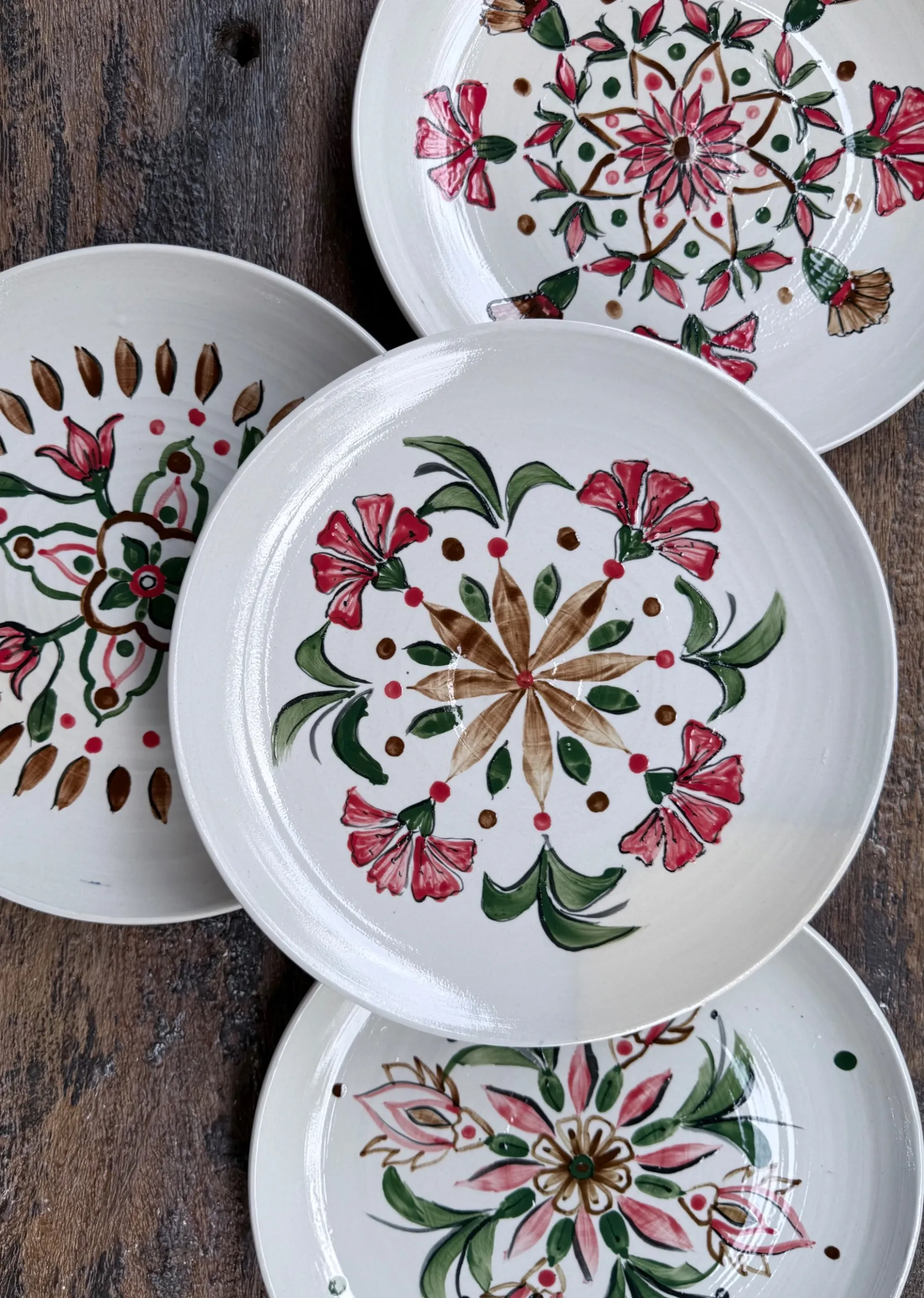 Four white ceramic plates featuring hand-painted red, pink, and green floral patterns on a dark wooden background.